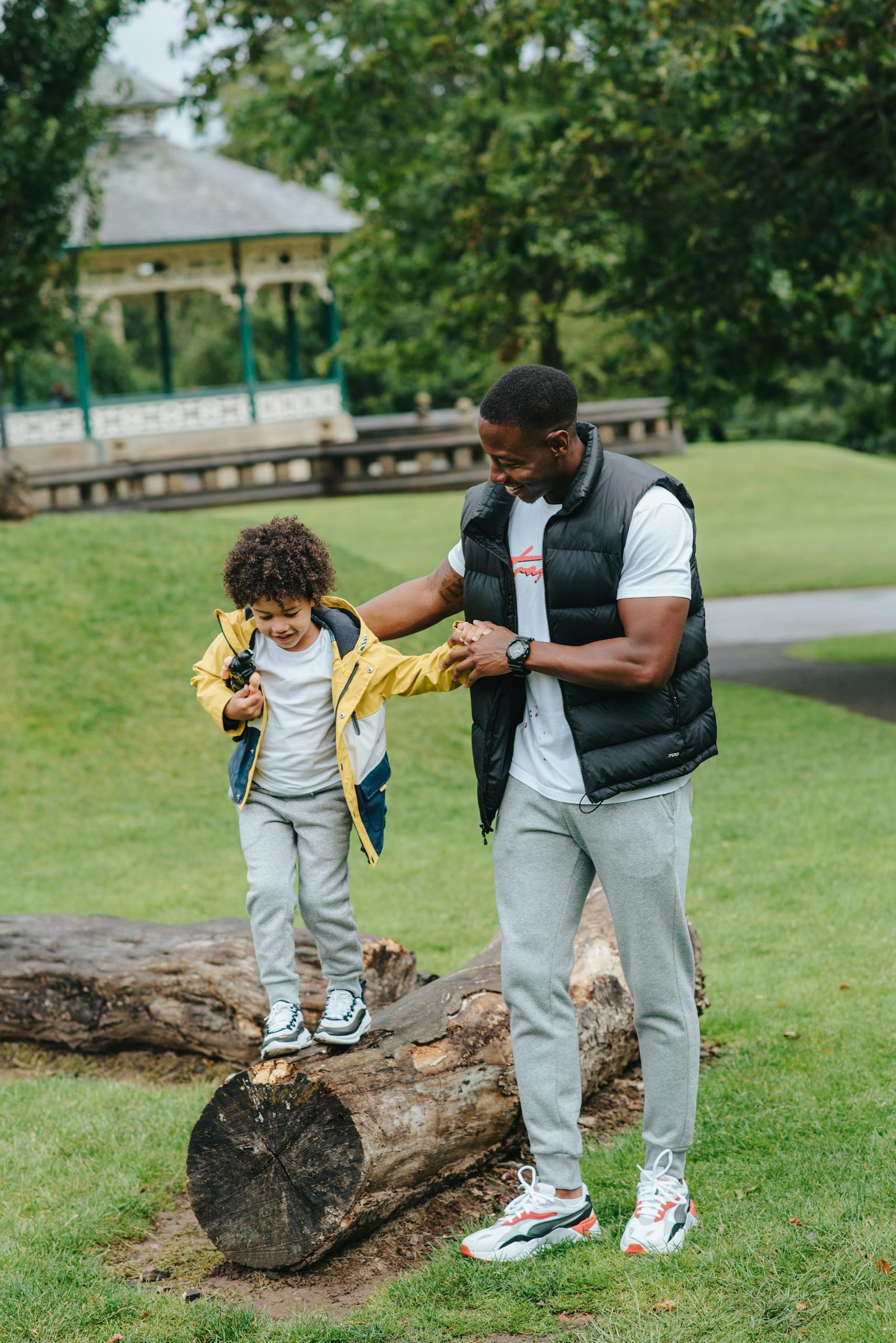 Full body of cheerful African American man holding hands of little boy while balancing on beam in park during weekend